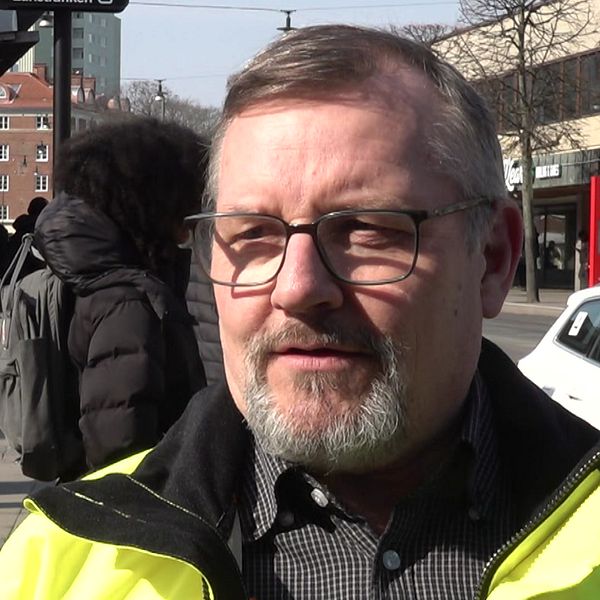 En person i gul reflexjacka vid busshållplats, passerande människor i bakgrunden.