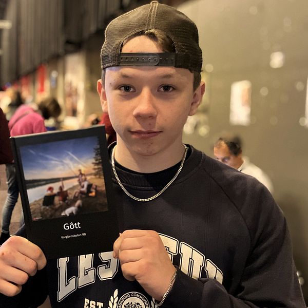 En person håller upp en bok med titeln ”Gött”, som har en bild av en strand på omslaget. I bakgrunden syns en folksamling på ett evenemang.