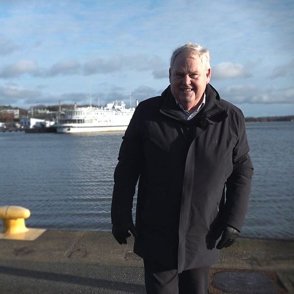 En person i svart jacka går längs en strandpromenad vid en hamn, med båtar som ligger förankrade i bakgrunden. Himlen är delvis molnig och vattnet verkar stilla.
