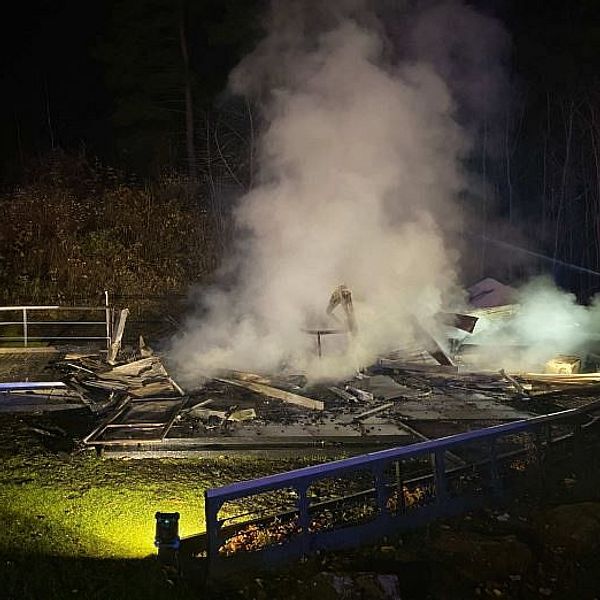 Rök stiger från resterna av en nerbrunnen byggnad i en skogsmiljö under natten. Brända träbjälkar och plåt ligger utspritt på marken, medan rök och dimma skingras i luften. En ljuskälla kastar sken på vraket.