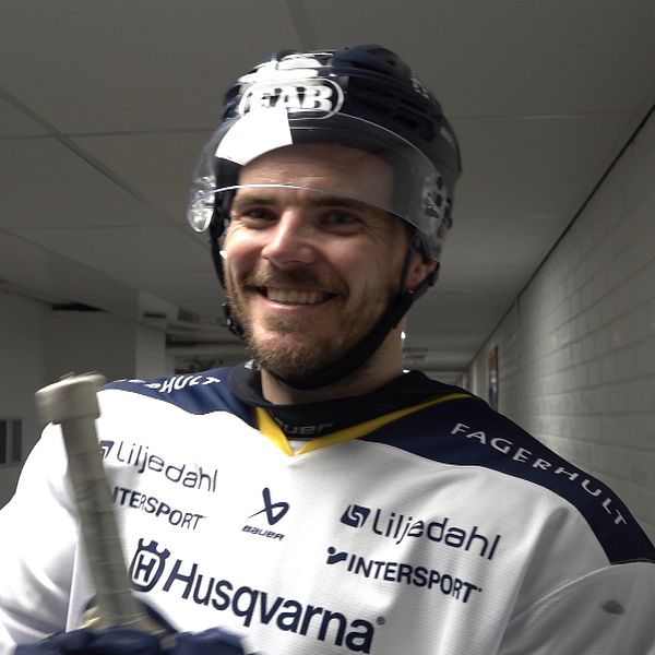 Oscar Engsund i en omklädningshall håller en klubba och en annan utrustning. Han är klädd i en tröja med sponsormärken och en hjälm, vilket antyder att han förbereder sig för en match eller träning. Bakgrunden är neutralt grå med belysning längs korridoren.