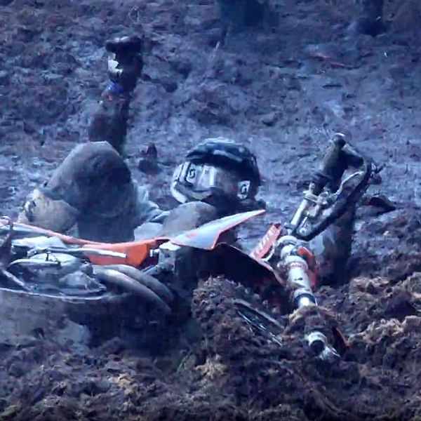 En motocrossförare ligger på marken efter att ha fallit av sin motorcykel, som är delvis nedsänkt i leran. Föraren är klädd i skyddsutrustning och lyfter en hand i luften, vilket ger en signal om att han fortfarande är vid medvetande trots olyckan. Området runt omkring är lerigt och blött.