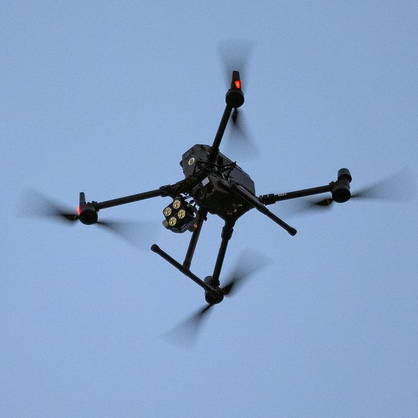 En svart drönare med roterande propellrar sväva i en klar himmel, synlig med tre av sina fyra ben och en kamera under kroppen.