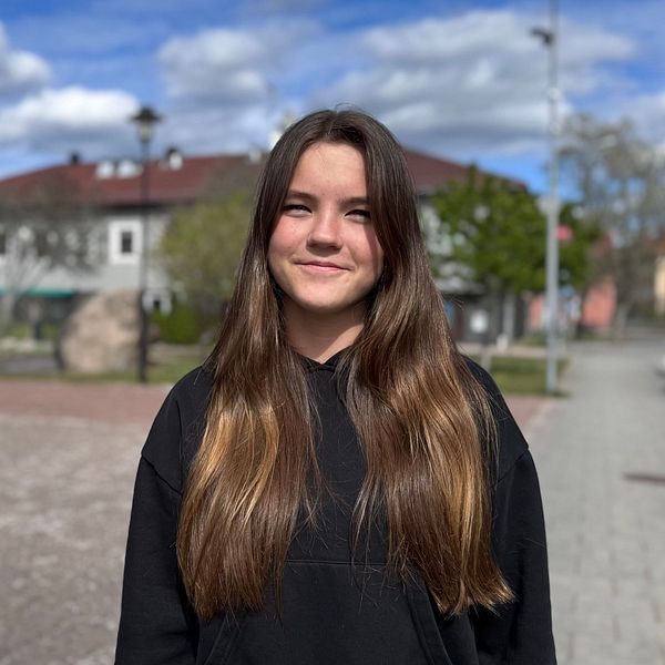 En person med långt brunt hår står på ett torg med byggnader och bilar i bakgrunden.