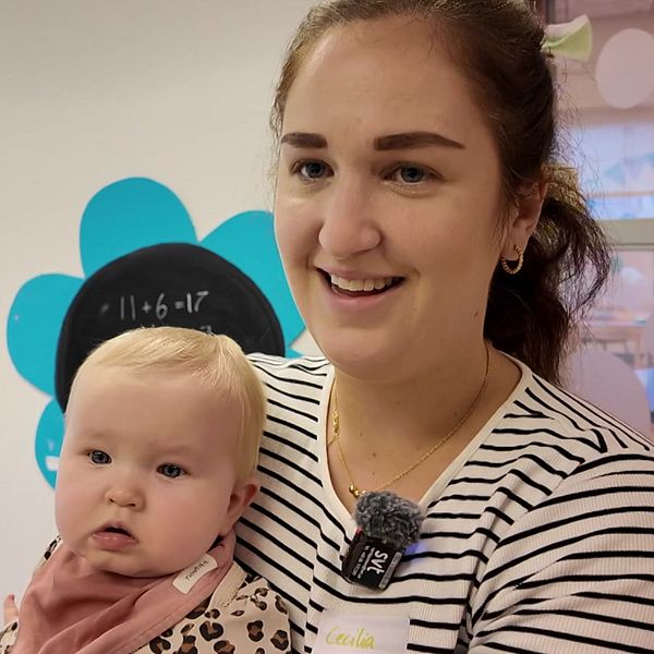 En kvinna håller ett litet barn i en förskolemiljö med färgglada blommor på väggarna. I bakgrunden finns tavlor i svart som kan användas för undervisning. Atmosfären verkar varm och inbjudande, vilket tyder på en lärorik och trygg plats för barn.