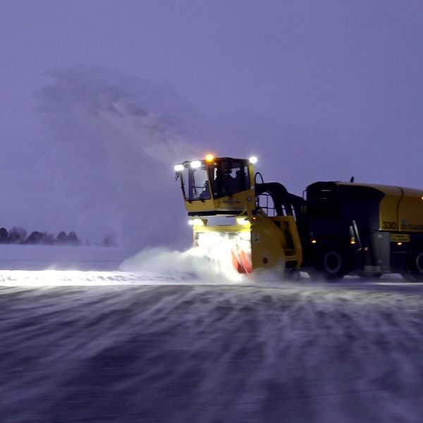 Snöröjare i aktion på en snötäckt flygplats under skymning, med kraftfulla lampor som lyser upp arbetsområdet medan snön blåser bort från banan. I bakgrunden syns en byggnad som kan vara en flygkontroll.