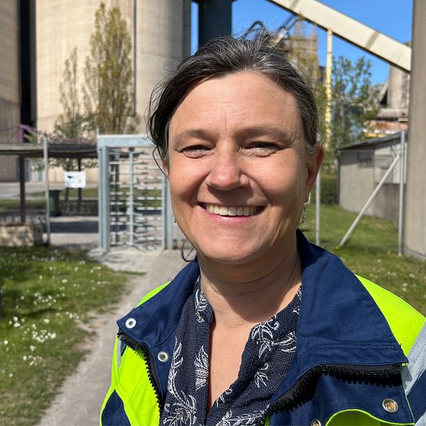 Leende person med reflexjacka utomhus vid industrimiljö med silos och grön gräsmatta.