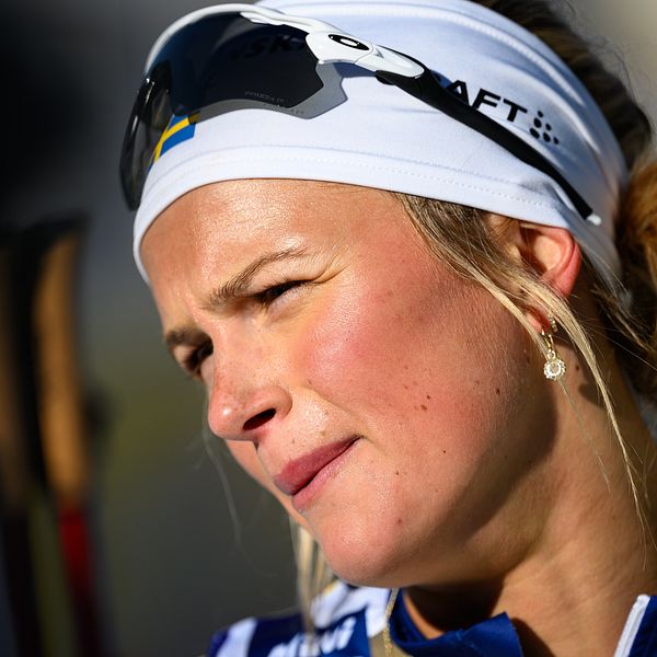 En person med en vit skidpannband och solglasögon står i profil. Personen bär skidutrustning som syns i bakgrunden.