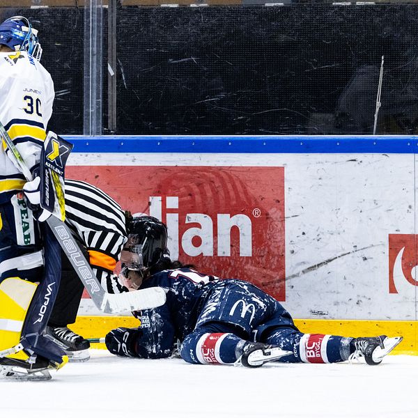 En ishockeyspelare ligger på isen medan en domare kontrollerar spelaren. En annan spelare står i närheten, vilket tyder på en incident på isen. Det finns en ishockeymål i bakgrunden.