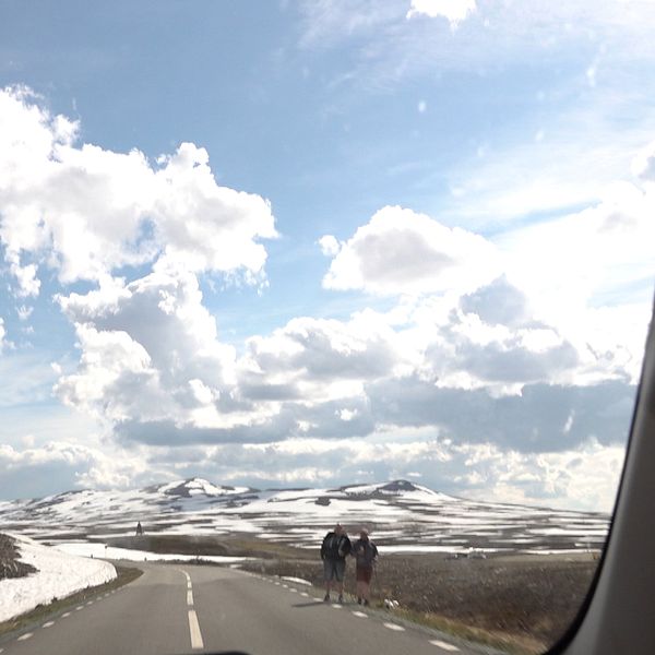 Landsväg genom fjällandskap med snö, två vandrare ses längre fram på vägen.