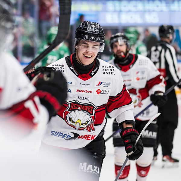 Ishockeyspelare i vita och röda tröjor på isen under en match. Domaren syns bakom.