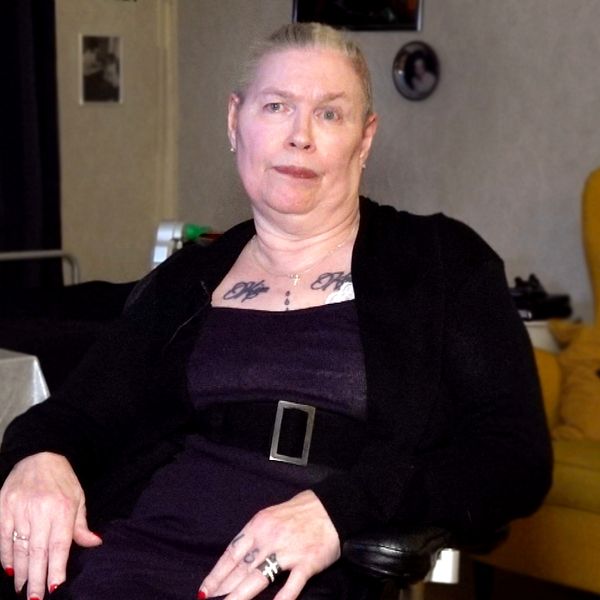En person sitter avslappnat i en rullstol, klädd i en svart jacka över en lila klänning. I bakgrunden syns en mysig inredning med mörka gardiner och en gul soffa. Bredvid personen finns ett bord med några tända ljus och dekorationer, vilket skapar en varm atmosfär.