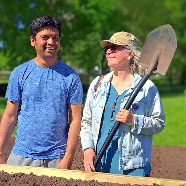 Två personer, en man med blå tröja som skrattar och en kvinna med spade, står framför odlingslåda