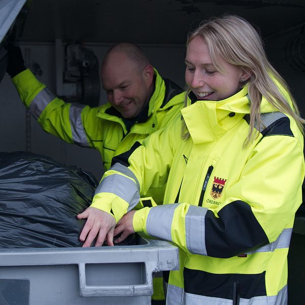 Två personer i neonfärgade arbetsjackor är i färd med att tömma en soptunna, där en stor svart sopsäck syns. Den ena personen sträcker sig in i tunnan för att hantera avfallet.