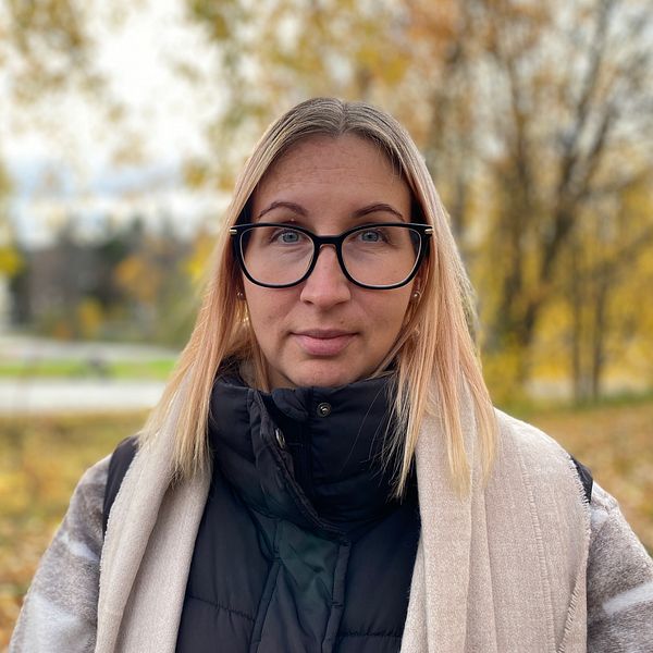 En person står i en skogsmiljö med höstfärger. Bakgrunden visar träd med gula och gröna löv samt en väg som sträcker sig bortom. Personen är klädd i en svart jacka och en ljus sjal, vilket ger ett avslappnat intryck i den naturnära omgivningen.