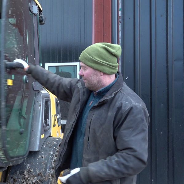 En person står vid en maskin och öppnar dörren. Maskinen har smutsiga ytor, vilket tyder på att den har använts i ett jord- eller byggarbetsprojekt. I bakgrunden syns en vägg av svart plåt, vilket ger en industriell känsla.