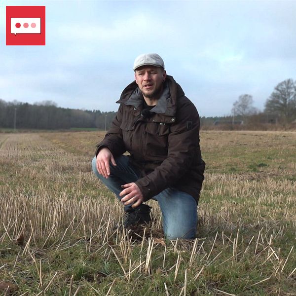 En person klädd i mörk jacka och jeans sitter på huk i ett öppet fält med kortklippt gräs eller stubb. I bakgrunden syns en skogskant och en molnig himmel.