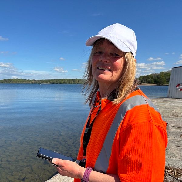 Person i orange arbetskläder vid sjö, håller en mobiltelefon
