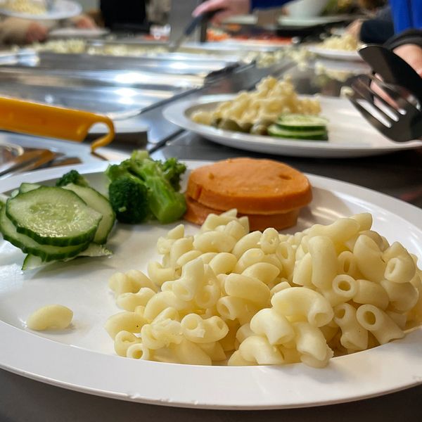 En tallrik med pasta, broccoli, gurkskivor och en bit korv, placerad på en serveringsdisk i en matsal. I bakgrunden syns andra maträtter och personer som serverar sig själva.