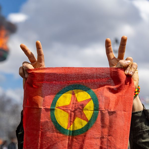 Person håller PKK-flagga och gör segertecken, brinnande fackla i bakgrunden.