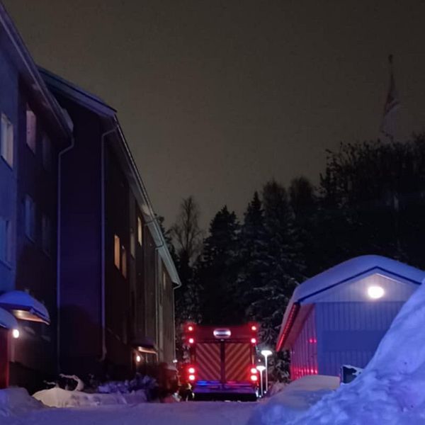Nattbild av en bostadsbyggnad med snö på marken, där en brandbil med lampor som blinkar står parkerad intill.