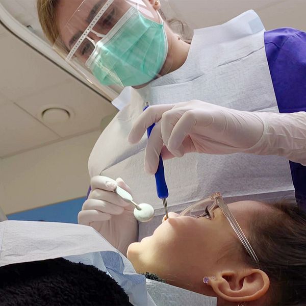 En tandvårdspersonal som arbetar med en patient under en tandbehandling. Tandläkaren håller ett instrument och fokuserar på patientens mun, medan det finns olika verktyg synliga i arbetsområdet. Patienten ligger på en behandlingsstol, och säkerhetsåtgärder som skyddande kläder används.