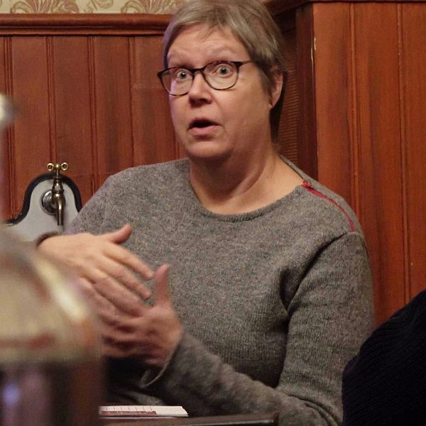 En kvinna i grå tröja och glasögon pratar vid ett konferensbord.