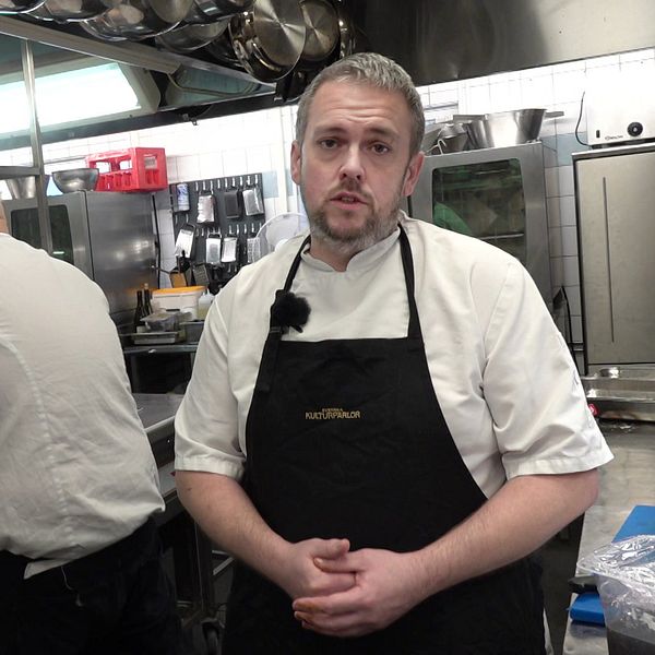 Kökschefen Andreas Eriksson står i köket och tittar mot kameran. I bakgrunden syns en kock steka kött.
