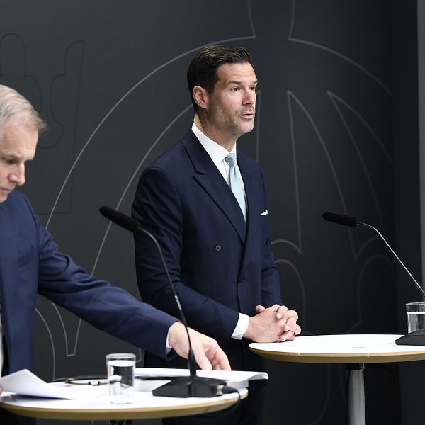 Två män i kostym står vid varsitt podium under en presskonferens.
