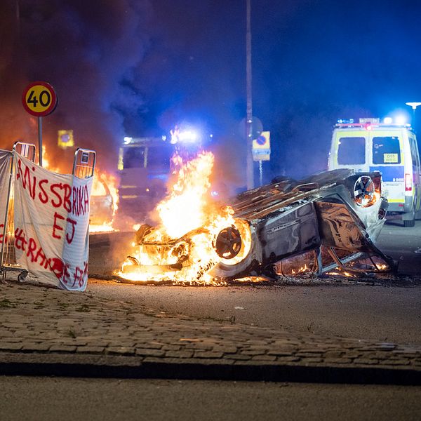 Bild från upploppet i Rosengård efter koranbränningarna i Malmö. Till vänster banderoll med texten: ”Nej till koranbränningar”, bredvid en brinnande bil och polisbil.