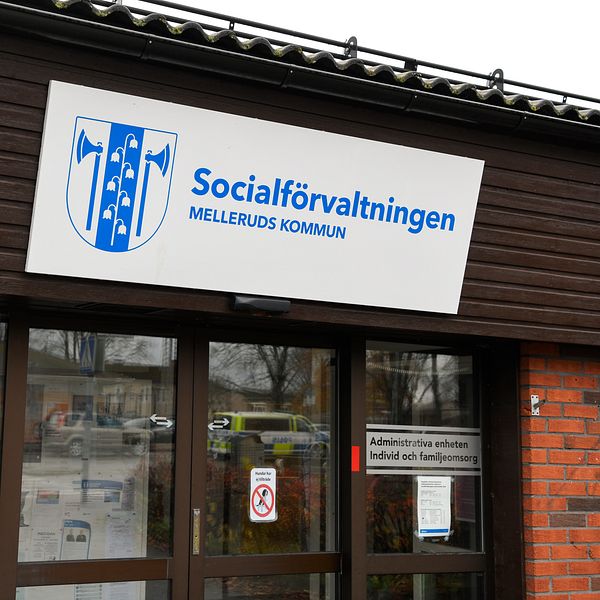 Entrén till Socialförvaltningen i Melleruds kommun, med en skylt som tydligt visar namnet på institutionen. Flera informationsskyltar och dokument är synliga på fönstren. Byggnaden har en tegelvägg och ett platt tak, i en urban miljö.