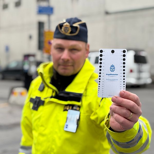 Polis i gul jacka håller upp ett polisdokument på en gata med bilar i bakgrunden.
