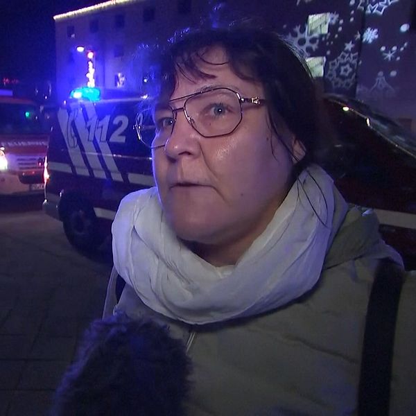 En person står på gatan med en vit scarf, medan räddningstjänstens fordon med blåljus syns i bakgrunden. Nattlig stadsmiljö med lysande dekorationer och olika ljus.