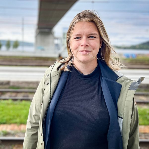 En person står framför en tågstation med järnvägsspår och en bro i bakgrunden. Det är en molnig himmel och landskapet är grönt med några träd i fjärran. Personen bär en ljusgrön jacka över en svart tröja. Linnéa Granqvist har en namninsamling om att bevara nattåg.