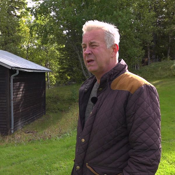 En äldre man i mörklila jacka står framför ett litet hus