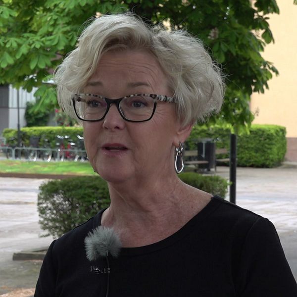 Kvinna intervjuas utomhus i ett bostadsområde med träd och byggnader i bakgrunden.