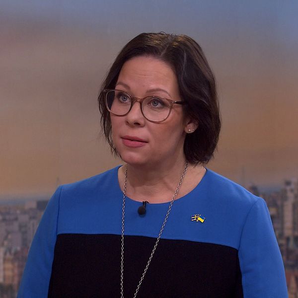 Maria Malmer Stenergard står framträdande i en blå och svart klänning, med ett svenskt flaggmärke synligt. Bakgrunden visar en stadsutsikt med byggnader och en ljus himmel.