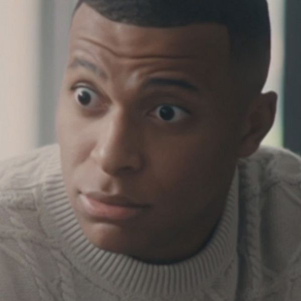 Kylian Mbappé i en vit, stickad tröja sitter på en soffa med en koncentrerad min. Bakgrunden är suddig och ljus, vilket skapar en mjuk och lugn atmosfär.