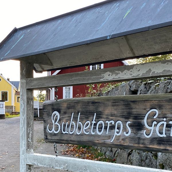 Skylt på Bubbetorps gård med vit text, i bakgrunden ett rött och ett gult hus samt ett höstfärgat träd.