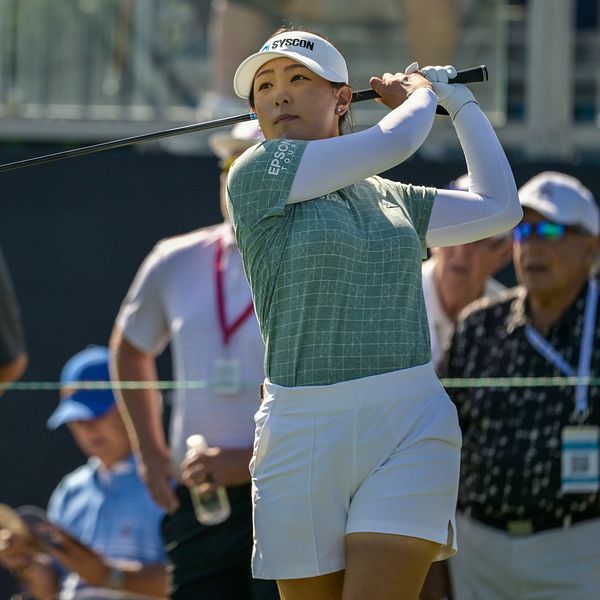 En golfspelare svingar sin driver på en golfbana, omgiven av åskådare som följer spelet. Det är en solig dag, och deltagarna är klädda i lämpliga golfkläder. I bakgrunden syns en del av golfbanan tillsammans med andra personer som är engagerade i spelet.