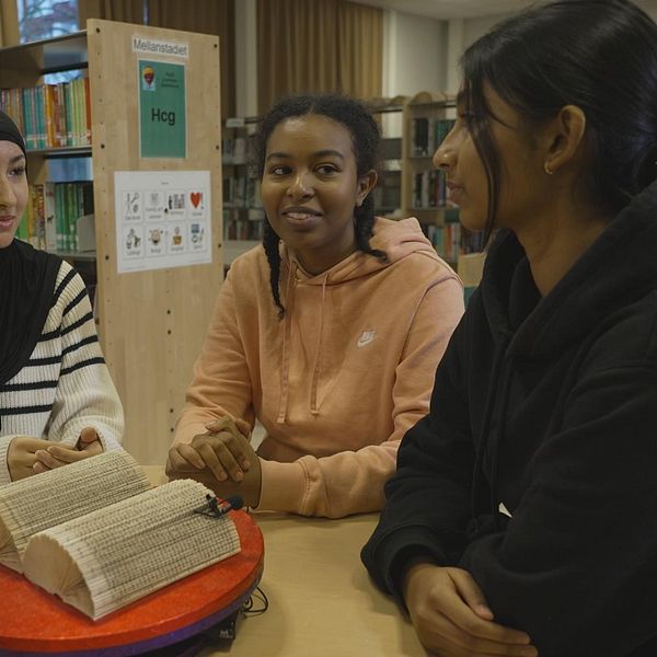 Grupp med fyra personer som sitter runt ett bord i en biblioteksmiljö. På bordet finns en stor bok, och deltagarna diskuterar eller interagerar.