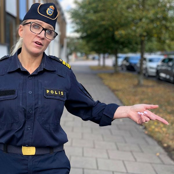 En polis i uniform står på en gata och pekar med ena handen, med träd och parkerade bilar i bakgrunden. Tina Anderberg. Våld i nära relationer.