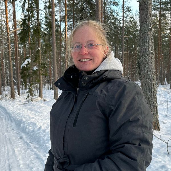 En kvinna klädd i en svart vinterjacka står på en väg i skogen som är täckt av snö. Hon tittar in mot kameran och ler.