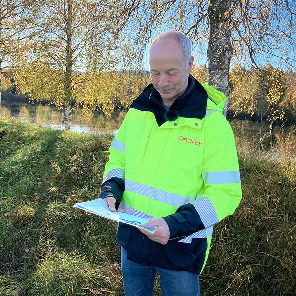 En person i en högsynlighetsjacka står vid en grön gräsyta nära en vattenspegel, med en vall och träd i bakgrunden. Personen håller i en karta.
