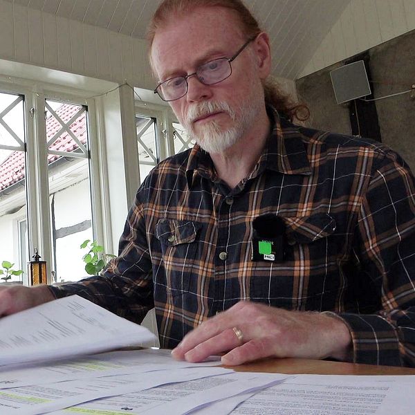 Paul Carlbark i rutig skjorta sitter vid ett bord med flera dokument framför sig. Rummet har stora fönster som släpper in naturligt ljus, och det finns inomhusväxter synliga i bakgrunden. På bordet står också ett ljus i en hållare.