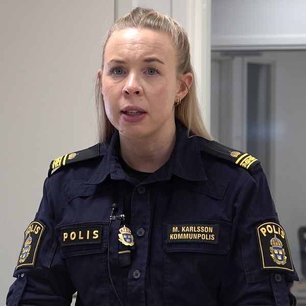 Kommunpolisen Maja Karlsson står och tittar mot kameran i sin mörkblåa polisuniform. Hon har blont hår, i bakgrunden är en vit dörr till vänster om henne och till höger ett fönster.
