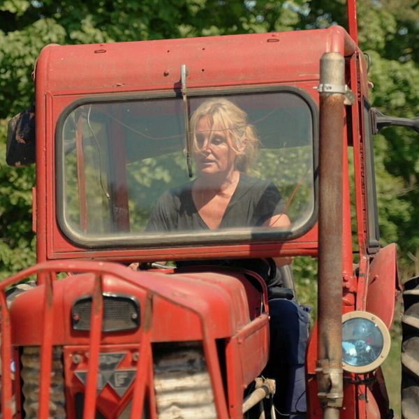 Kvinna kör röd traktor framför grönskande träd i lantlig miljö.