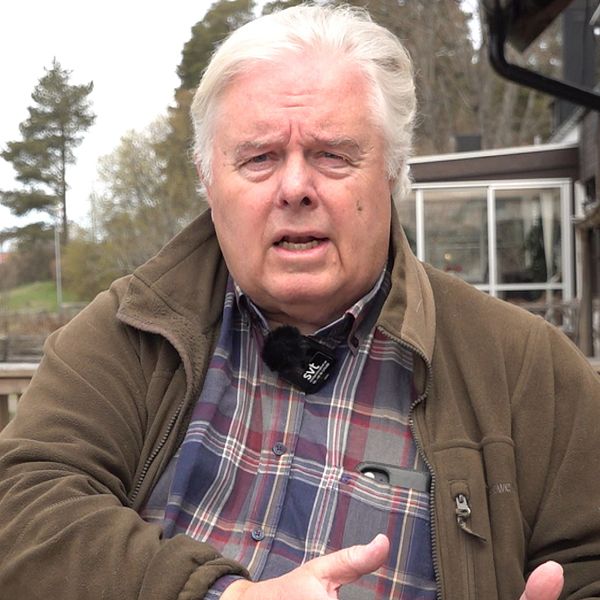En person i rutskjorta sitter utomhus vid ett trästaket, med en röd fågelholk i bakgrunden.