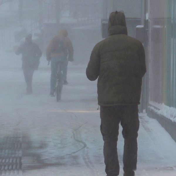 Mörk och snöig stadsmiljö med svag sikt på grund av kraftig snöblandat väder. I förgrunden går en person klädd i en mörk jacka, medan två andra personer, varav en på cykel, syns längre bort i en snöig landskap. Gatuskyltar och byggnader är delvis synliga genom snöverkan.