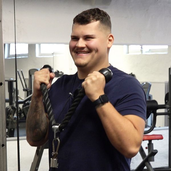 En ung man står på ett gym och tränar triceps. Han håller i ett träningsredskap och tittar in i en mobilkamera och ler.